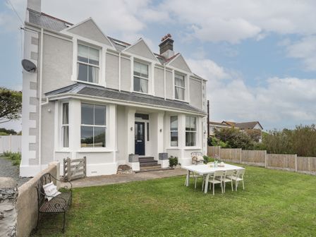 Exterior view of a two-story house with a front yard featuring a metal bench and a white outdoor dining table with chairs at 1 Tirionfa in Trearddur Bay