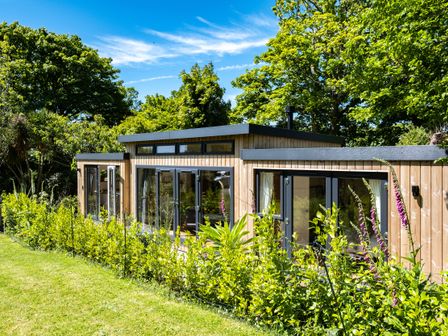A wooden single story house with large windows surrounded by green plants and trees at Chi Lowarth in Porthleven