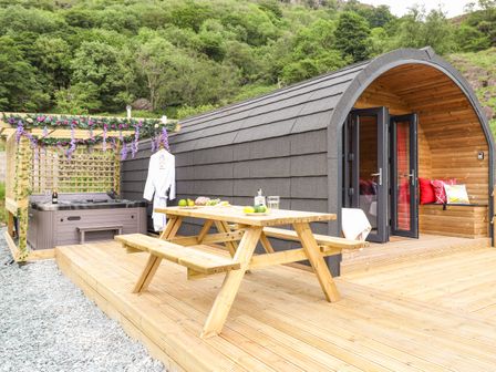 A wooden patio with a picnic table hot tub and arched cabin in a forested area at Hartsop Magic - Crossgate Luxury Glamping Hartsop near Glenridding