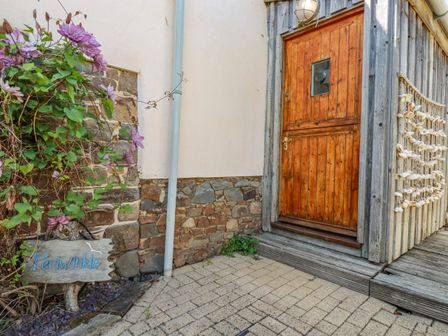 A wooden door entrance with stone wall and purple flowers beside a sign reading Periwinkle at Atlantic Highway near Bude