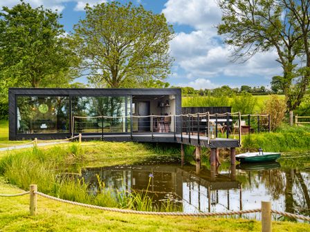 A modern black cabin with large glass windows beside a pond with a small boat and surrounded by green trees and grass at Rusty Boathouse in Llanarth