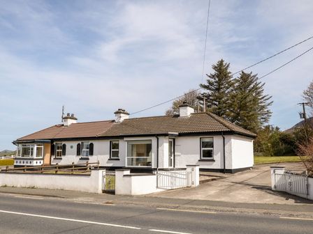 A white single-story house with a tiled roof and chimneys next to a paved driveway and trees at Bridgetown Cottage in Kerrykeel County Donegal