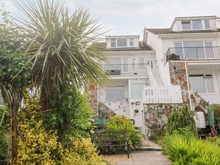 A multi-level house with a stone foundation and white exterior with stairs leading to balconies and a garden with plants and a bench at Flat 6 in Falmouth