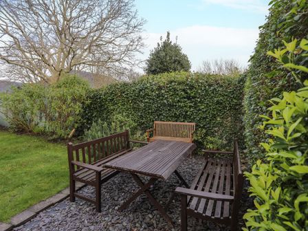 An outdoor seating area with a wooden table and three wooden benches surrounded by hedges and trees at Amelie Cottage in Beaumaris Anglesey