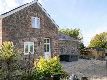 An outdoor view of a stone house with white windows and a door with a patio area and garden plants at Hope House Stables in Witheridge