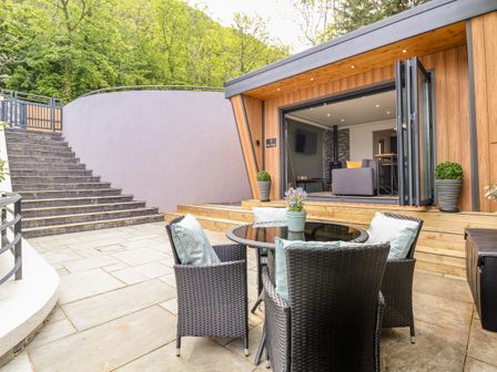 An outdoor patio with a round glass table and four wicker chairs with cushions in front of a modern wooden building with open folding doors at The Nest in Prestatyn