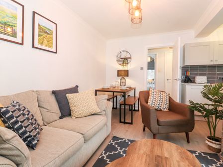 A living room with a beige sofa, a brown armchair, a wooden coffee table, a dining table with set dishes, and a kitchen area at Bluebell Hill in Bowness-On-Windermere