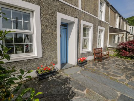A stone patio with a blue door two windows flower boxes and a wooden bench at 6 Glyn Terrace in Borth-Y-Gest near Porthmadog
