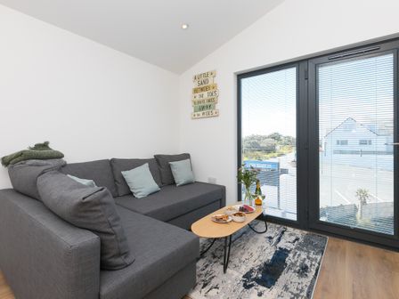 A living room with a gray sofa a wooden coffee table with snacks a glass door with blinds and a decorative sign on the wall at The Sandbox in Hayle