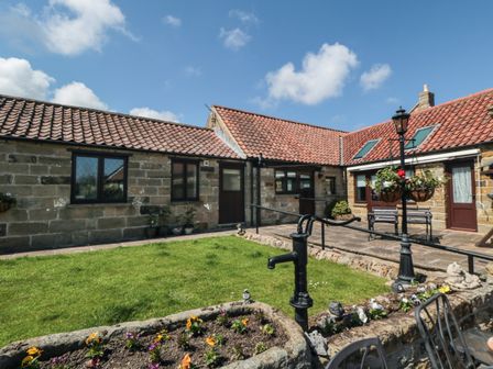 A courtyard with grass and flower beds surrounded by stone buildings with tiled roofs at Beaconsfield Cottage Burniston near Scalby