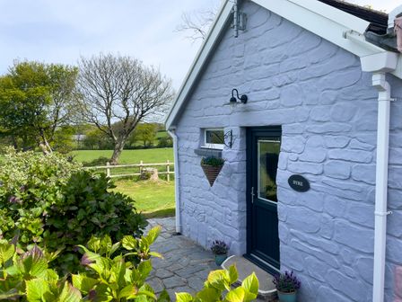 An exterior view of a stone building with a blue door and hanging flower basket next to a garden and trees at The Byre @ Canllefaes Penparc near Cardigan