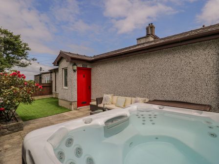 An outdoor area with a hot tub a sofa with pillows a red door and a small grassy patch at Bwthyn Pengwern in Saron near Caernarfon