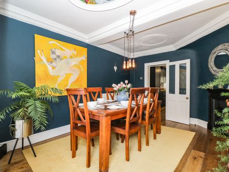 A dining room with a wooden table six chairs a yellow rug a plant and a painting on a blue wall at Cuckoo Cottage in Upton Upon Severn