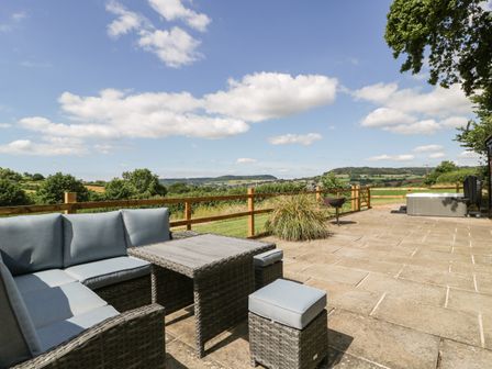 An outdoor patio with seating table a barbecue and a hot tub overlooking fields and hills at The Old Kennels in Harcombe near Sidford