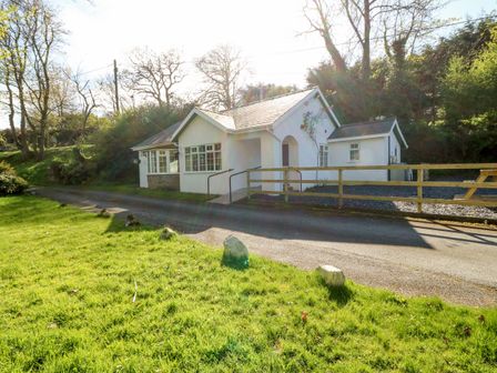A white cottage with a driveway and wooden fence surrounded by trees and grass at Mill House Cottage in Sarn Meyllteyrn