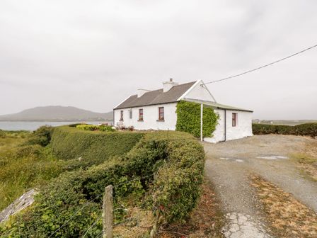 A white cottage with red window frames surrounded by hedges near a body of water at The White House in Roundstone County Galway