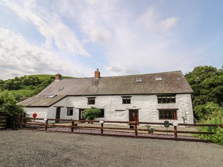 A white stone house with a sloped roof and multiple windows behind a wooden fence with hills and trees in the background at Penarth in Cregrina near Builth Wells
