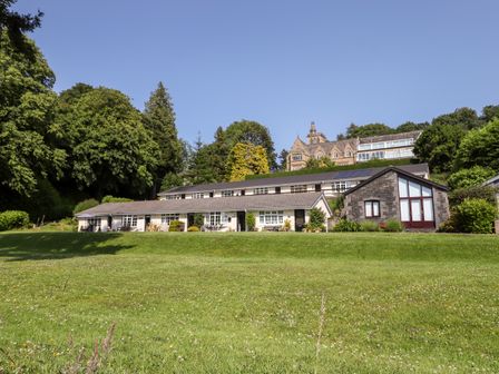 A green lawn with a row of white cottages and a stone building surrounded by trees at Ginger & Pickles in Windermere