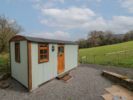 A small corrugated metal hut with wooden door and windows next to a gravel path and green field at Rabbit Hill Hut in Govilon