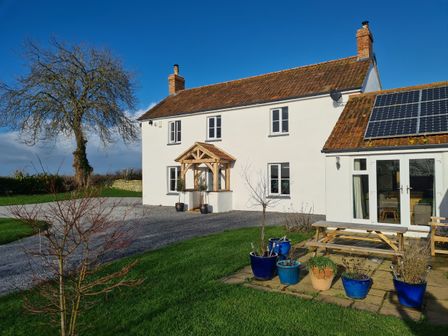 A white house with a gravel driveway and solar panels on the roof with potted plants and a wooden picnic table on a patio at Somerset House in Othery near Middlezoy