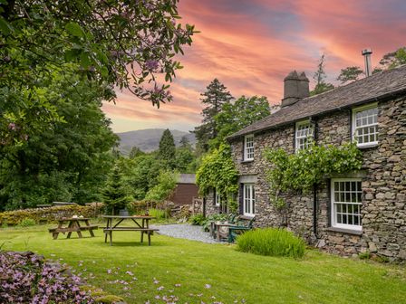 A stone cottage with windows and outdoor seating in a garden with trees under a sunset sky at Little How in Grasmere