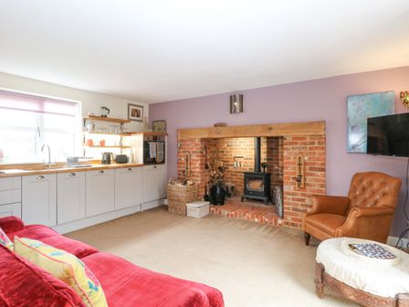 A living room with a kitchen area a red sofa a leather armchair a brick fireplace and a mounted television at Willow Cottage in Hickling near Catfield