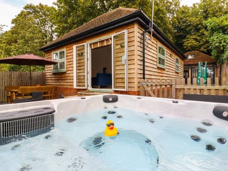 An outdoor hot tub with a rubber duck in front of a wooden cabin with open doors and outdoor seating with an umbrella at Dreamwood Lodge in Keston near Leaves Green