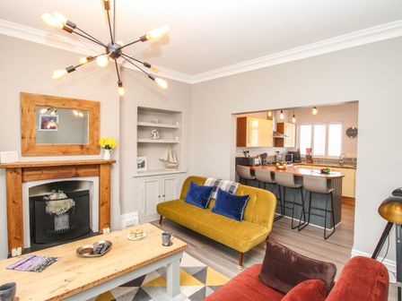 A living room with a yellow sofa, wooden coffee table, fireplace, and an open kitchen with bar stools at Swanage Bay in Swanage