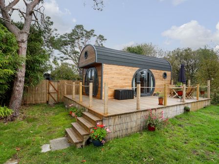 A wooden cabin with a deck surrounded by trees and grass at The Hideaway Pod in Rhoslan near Criccieth