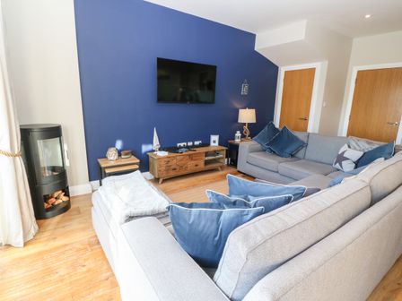 A living room with a grey sectional sofa and blue cushions a wooden TV stand with a TV and a wood stove at Sandy House in Rhosneigr