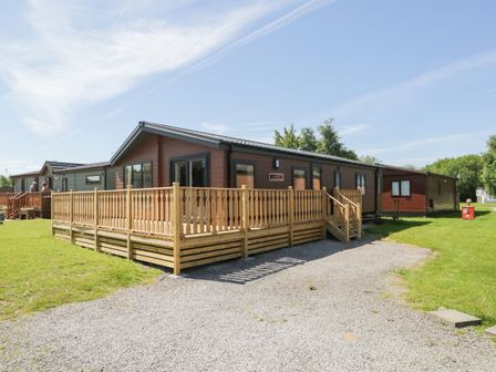 A wooden lodge with a fenced porch and gravel driveway on green grass under a blue sky at Parkers Place in Carnforth
