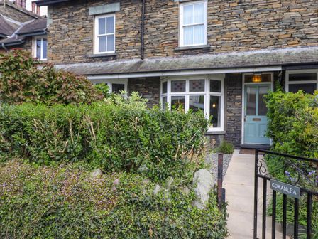 A stone house with a bay window and a blue door behind a hedge and a metal gate with the name Gowanlea in Ambleside