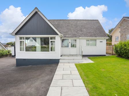 A white house with a dark roof and a paved walkway leading to the front door with grass on one side at Swn Y Mor in Benllech