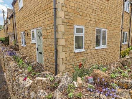 Exterior of a stone house with a green door and multiple windows surrounded by a stone wall with plants at The Cornerstone in Bourton-On-The-Water