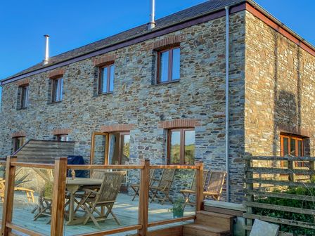 An outdoor wooden patio area with a table and chairs next to a stone house with multiple windows at Kestrel 19 Dartmouth Green Blackawton