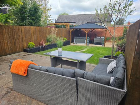 A backyard with outdoor seating, a glass table, green lawn, garden plants, a gazebo with hot tub and chairs at Rhif Saith in Ruthin