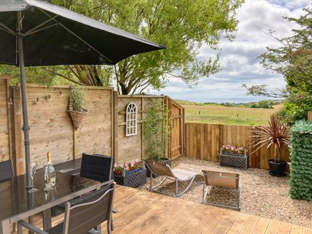 An outdoor patio with a table and chairs under an umbrella a wooden fence plants and lounge chairs at Little Honeynook in Bigbury
