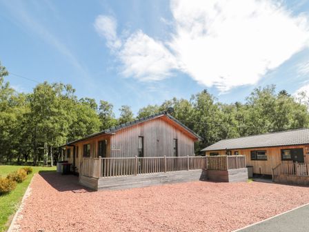 A wooden cabin with a fenced porch surrounded by trees and gravel in front at Lodge 32 Otterburn