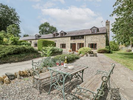 An outdoor seating area with wooden benches and table on gravel in front of a stone cottage house with multiple windows at Bullfinch Cottage in Kingsland