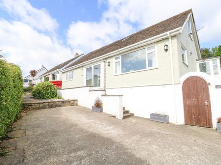 A driveway leading to a beige house with a wooden gate and shrubs at Treestumps in Benllech