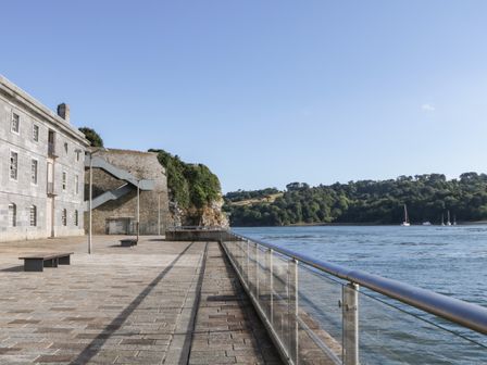 A waterfront promenade with benches and a stone building next to a river and wooded hills at Clarence Riverside Royal William Yard in Plymouth