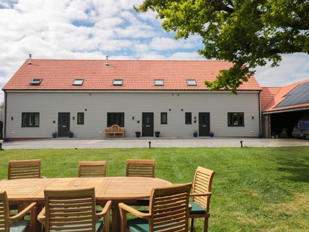 A wooden outdoor dining table with chairs on a lawn in front of a large building with red tiled roof at The Stables in Watchfield near Highbridge