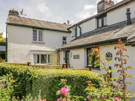 An exterior view of a white cottage with a sign that reads Anniversary Cottage surrounded by plants and bushes in Ambleside