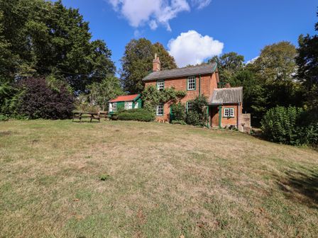 A brick house with green trim surrounded by trees and grass with picnic tables in the yard at Warren Cottage Bures near Assington