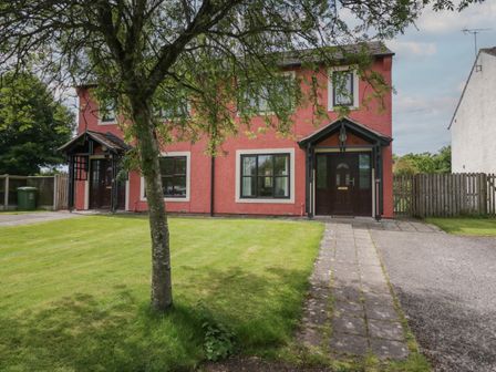A semi detached house with red exterior walls green lawn and tree in front at 9 Eamont Park in Penrith