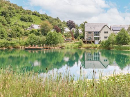 A house near a pond with a wooden dock and chairs surrounded by grass and trees at Tan Yr Allt in Dolanog near Meifod Powys