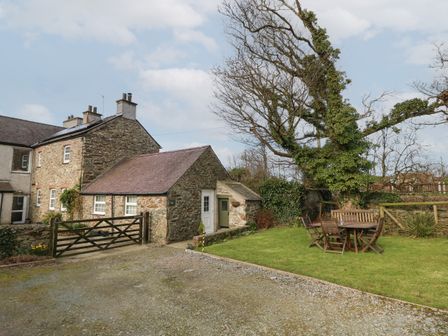 An outdoor area with a stone house, a gravel driveway, a wooden gate, a tree with ivy, and a wooden table with chairs on grass at Ty Gwenyn in Rhydwyn