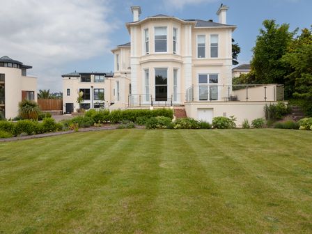 A large lawn in front of a cream-colored house with multiple windows and a glass-railed terrace at Rutland Heights in Torquay