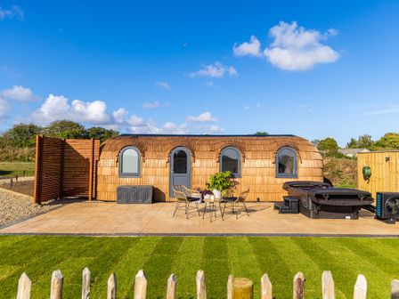 A small wooden cabin with arched windows and door on a paved patio with chairs plants and a hot tub at Andrus - Mayflower Meadow in Brandesburton