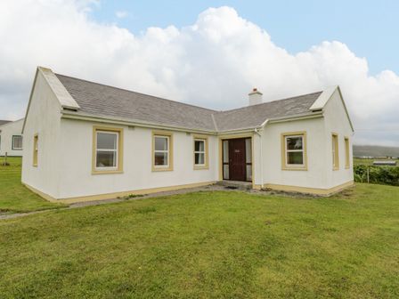 A single story house with white walls and a dark front door on a grassy lawn at 13 Trawmore Cottages in Keel County Mayo